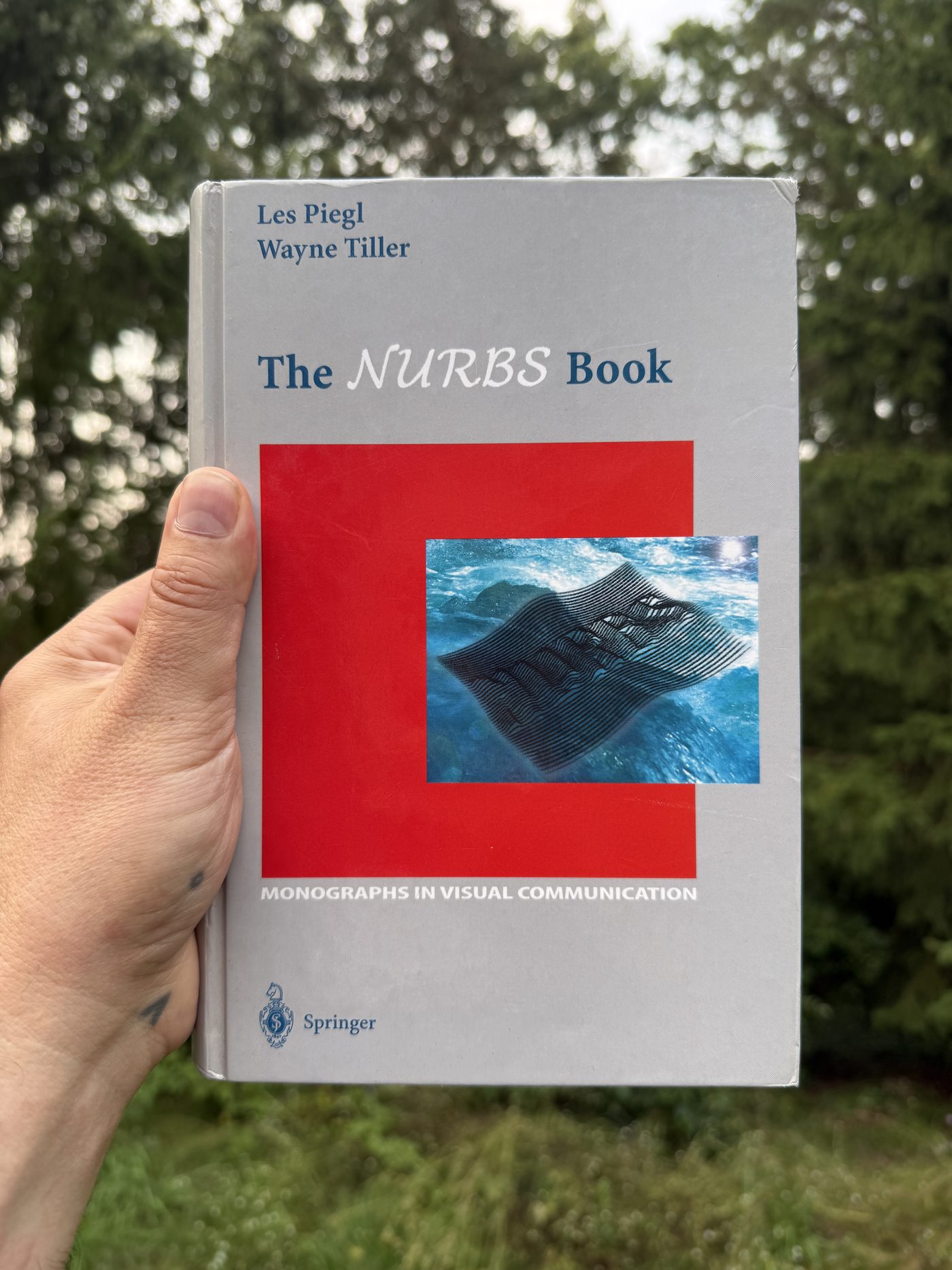 A hardback copy of The NURBS Book by Les Piegl and Wayne Tiller, held up outdoors against foliage.